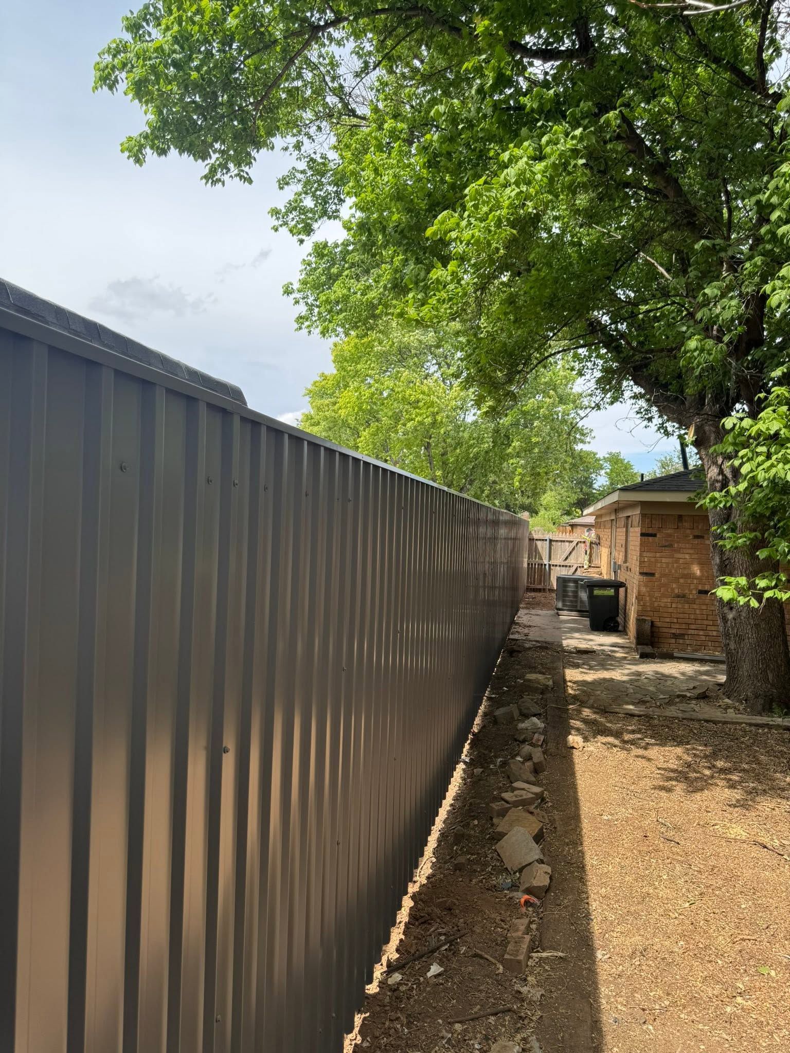Narrow yard beside a corrugated metal fence, with trees overhead and a small brick shed in the distance.