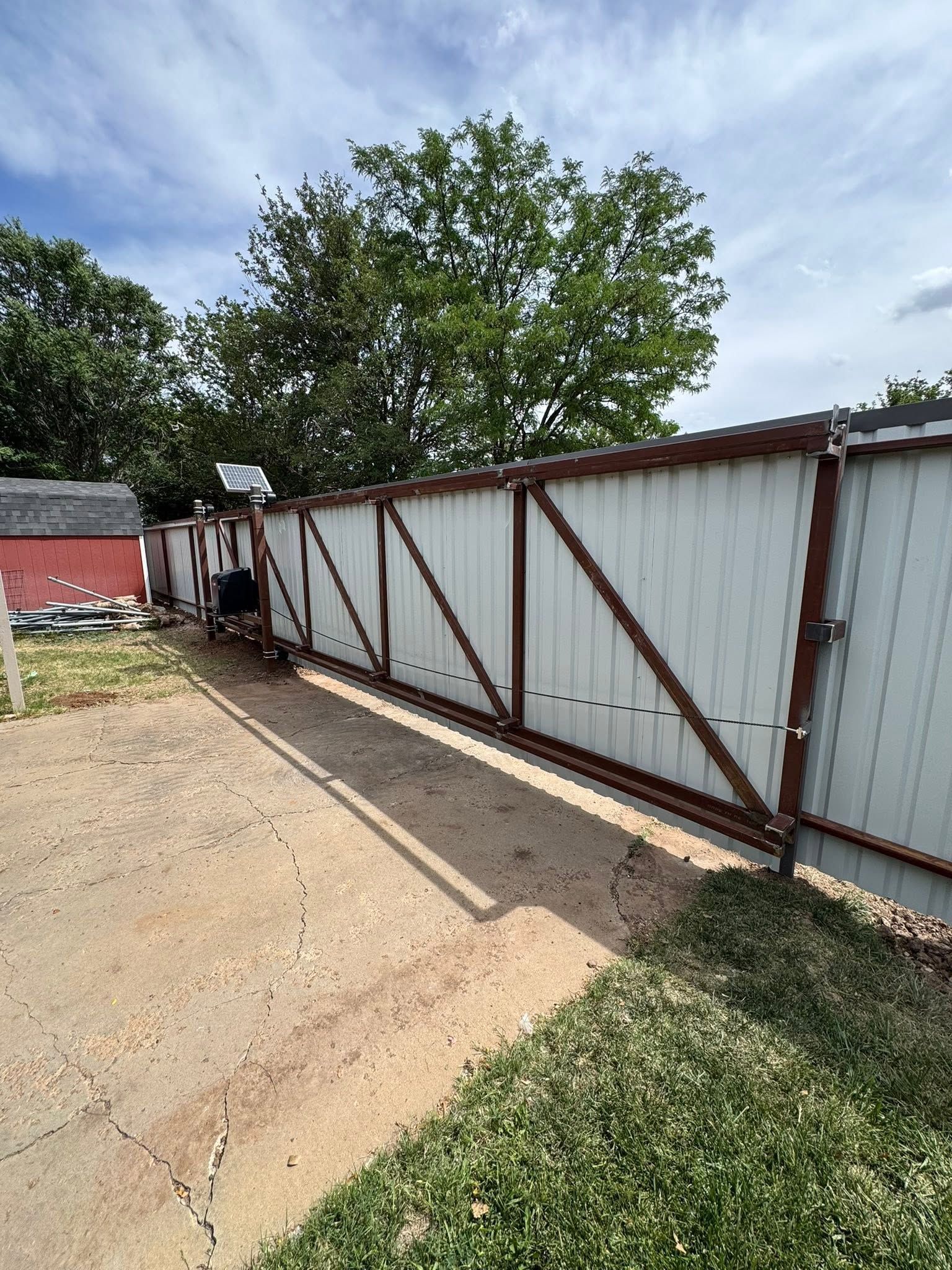 Long white metal sliding gate with brown supports beside a dirt driveway and grassy yard under a blue sky