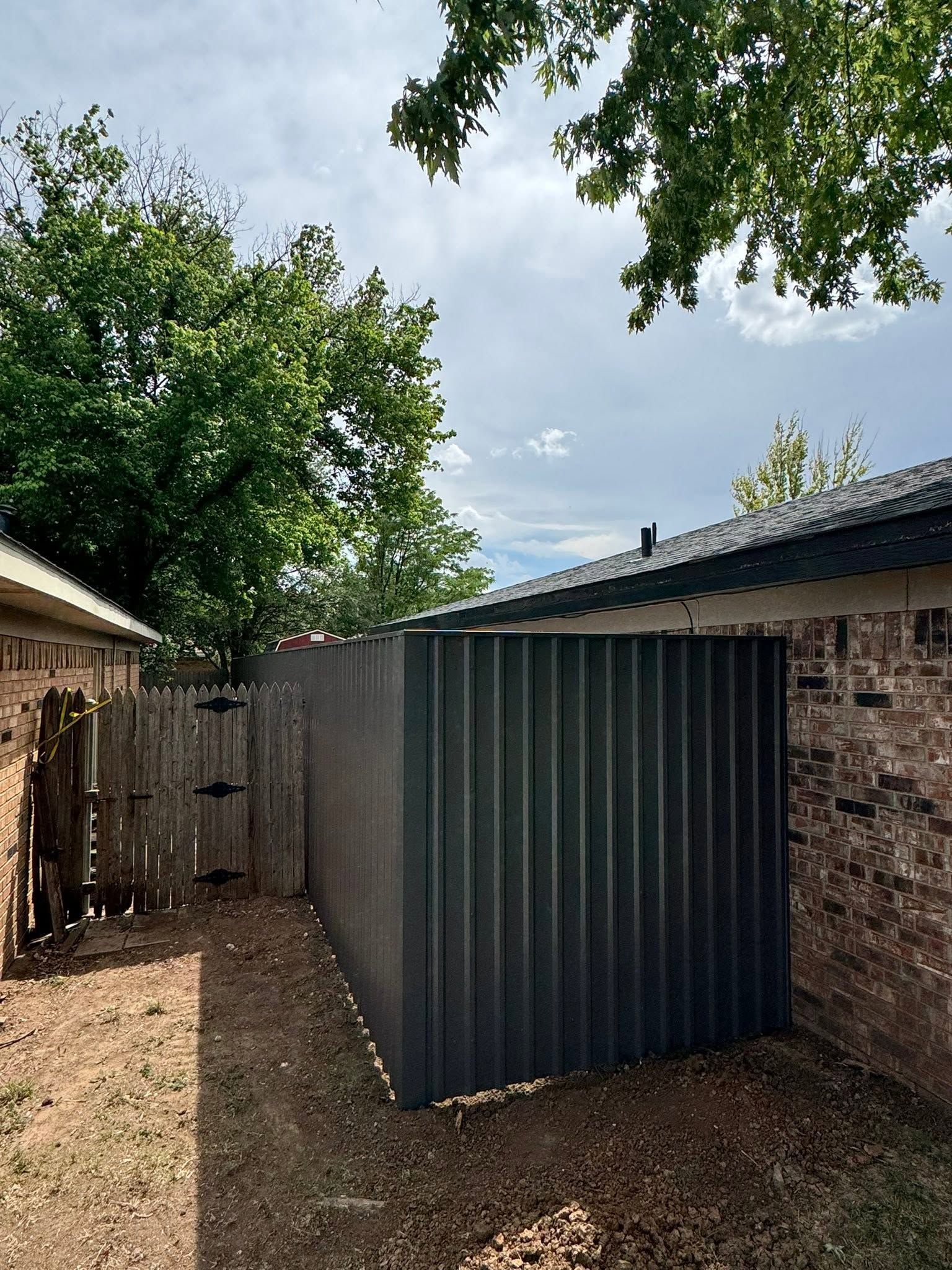 Backyard with a corrugated metal shed beside a brick house, surrounded by trees and a wooden fence.