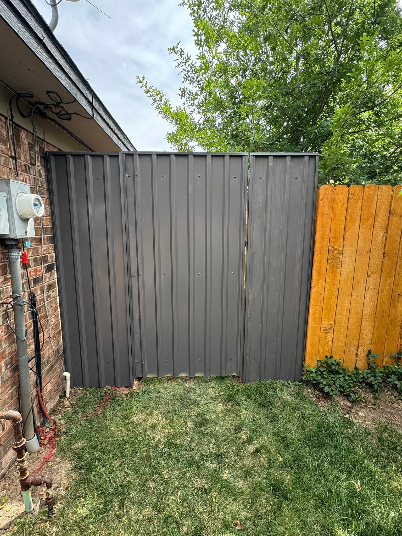 Gray metal gate in a grassy yard beside a brick wall and orange wooden fence.