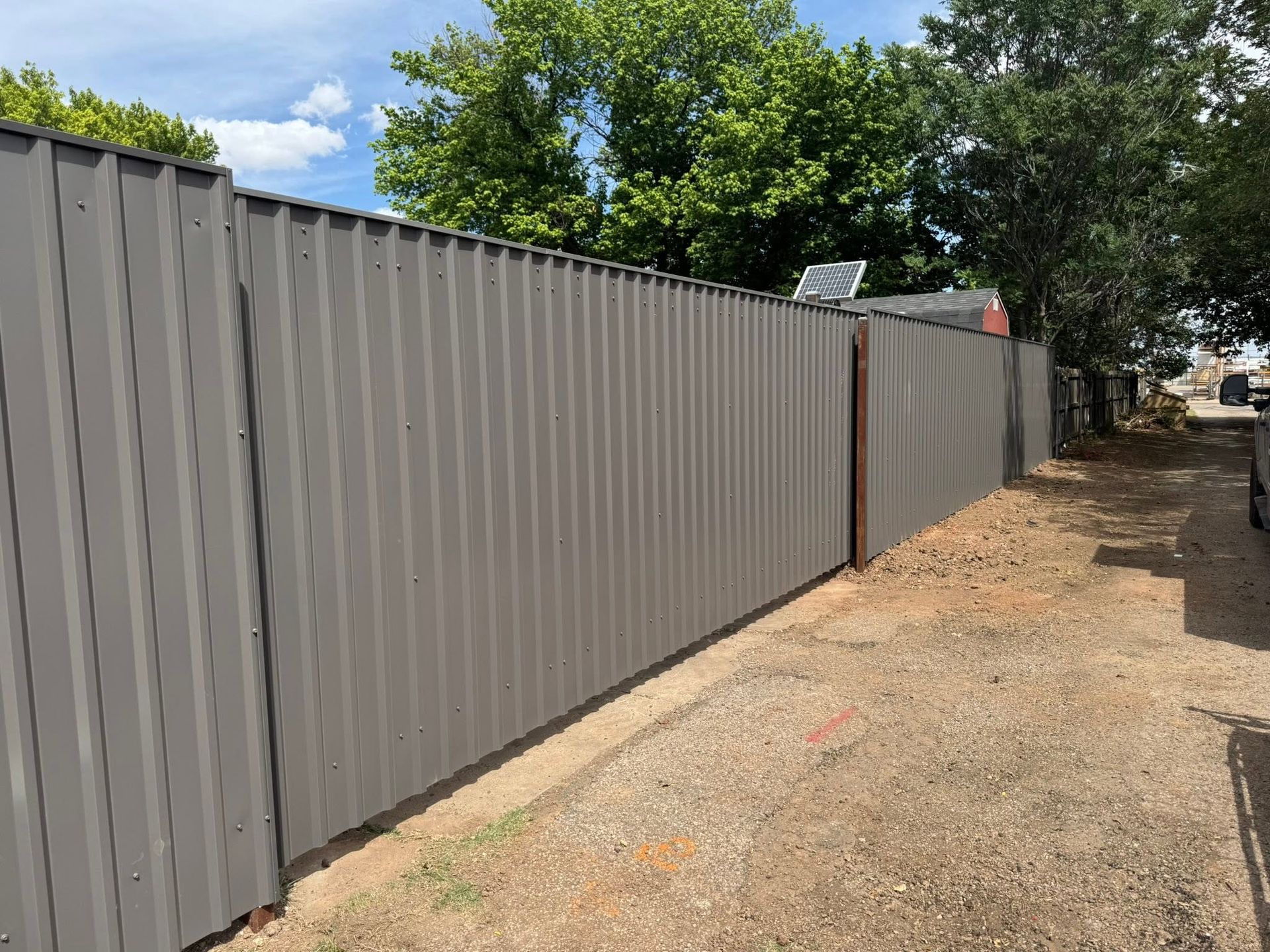 Gray corrugated metal fence along a dirt path beside trees and a blue sky