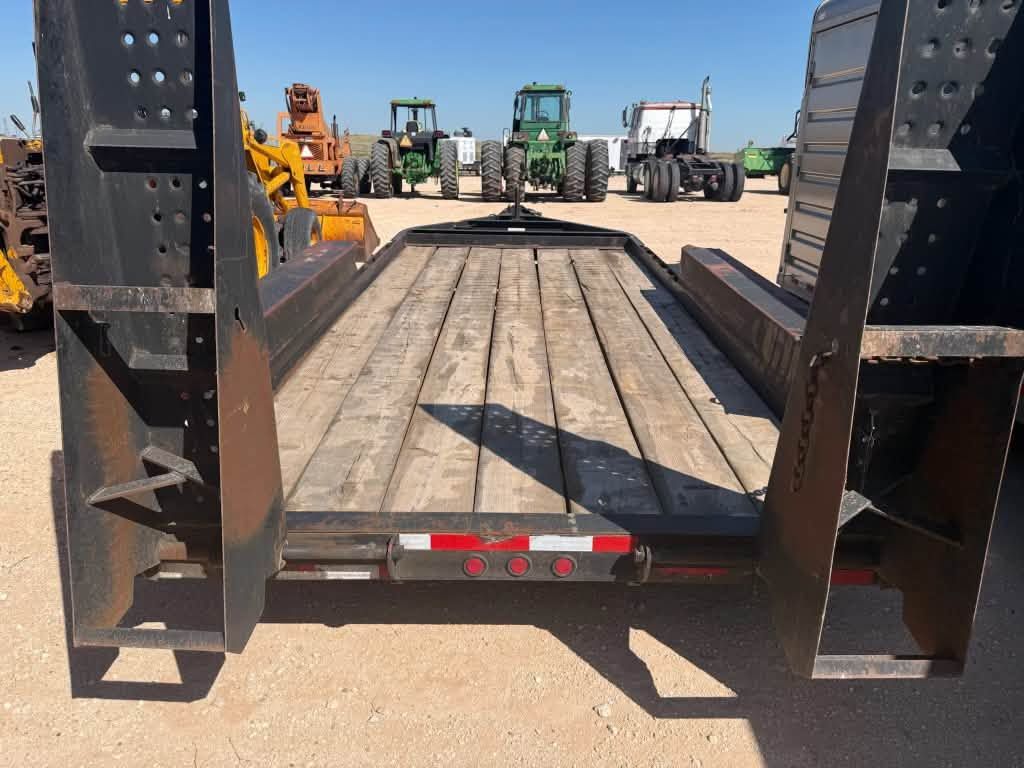 Empty flatbed trailer deck with black ramps, parked on gravel lot with farm equipment in the background