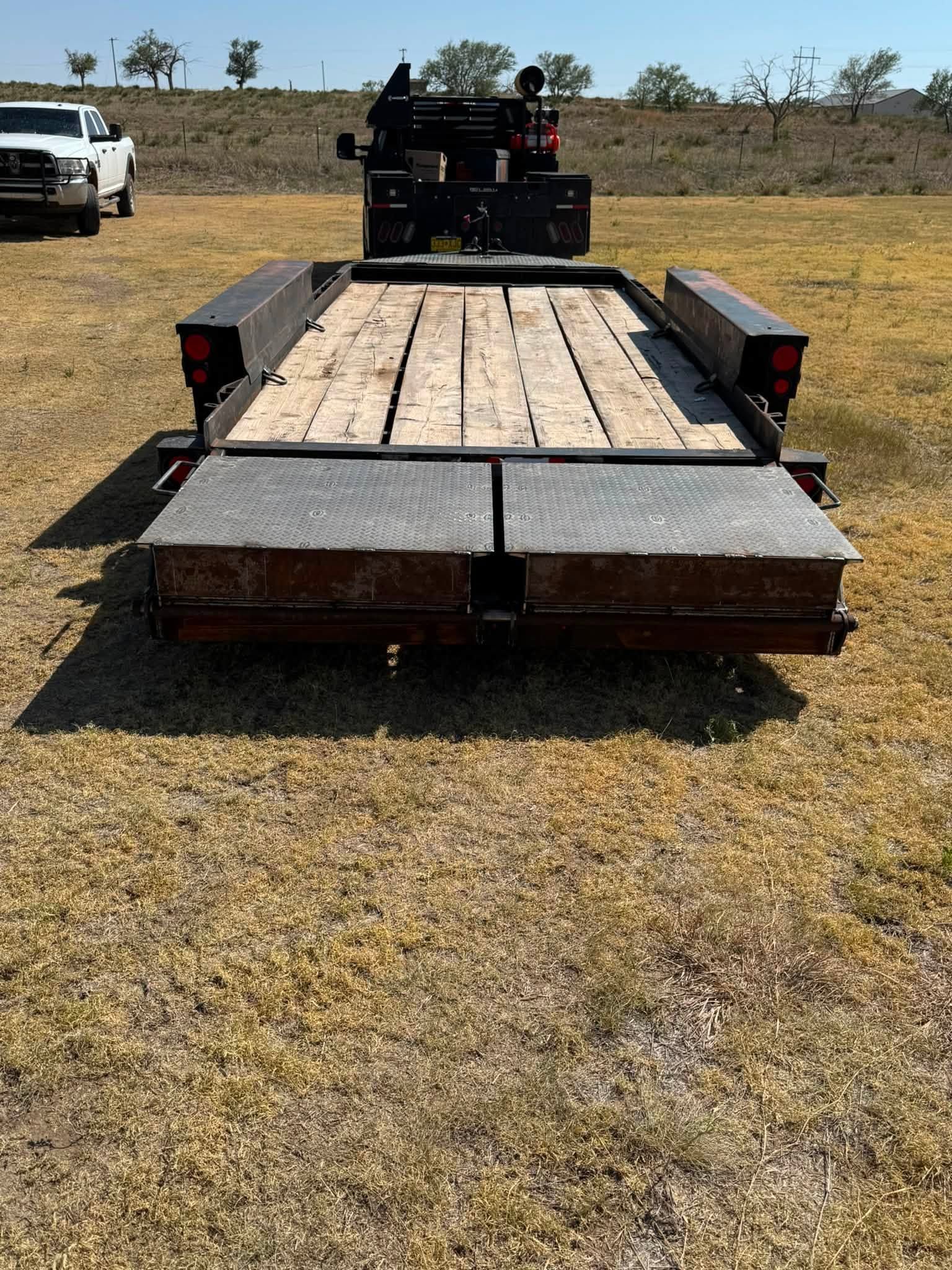 Flatbed trailer with wooden deck and metal ramps parked on a grassy field, with a truck in the background