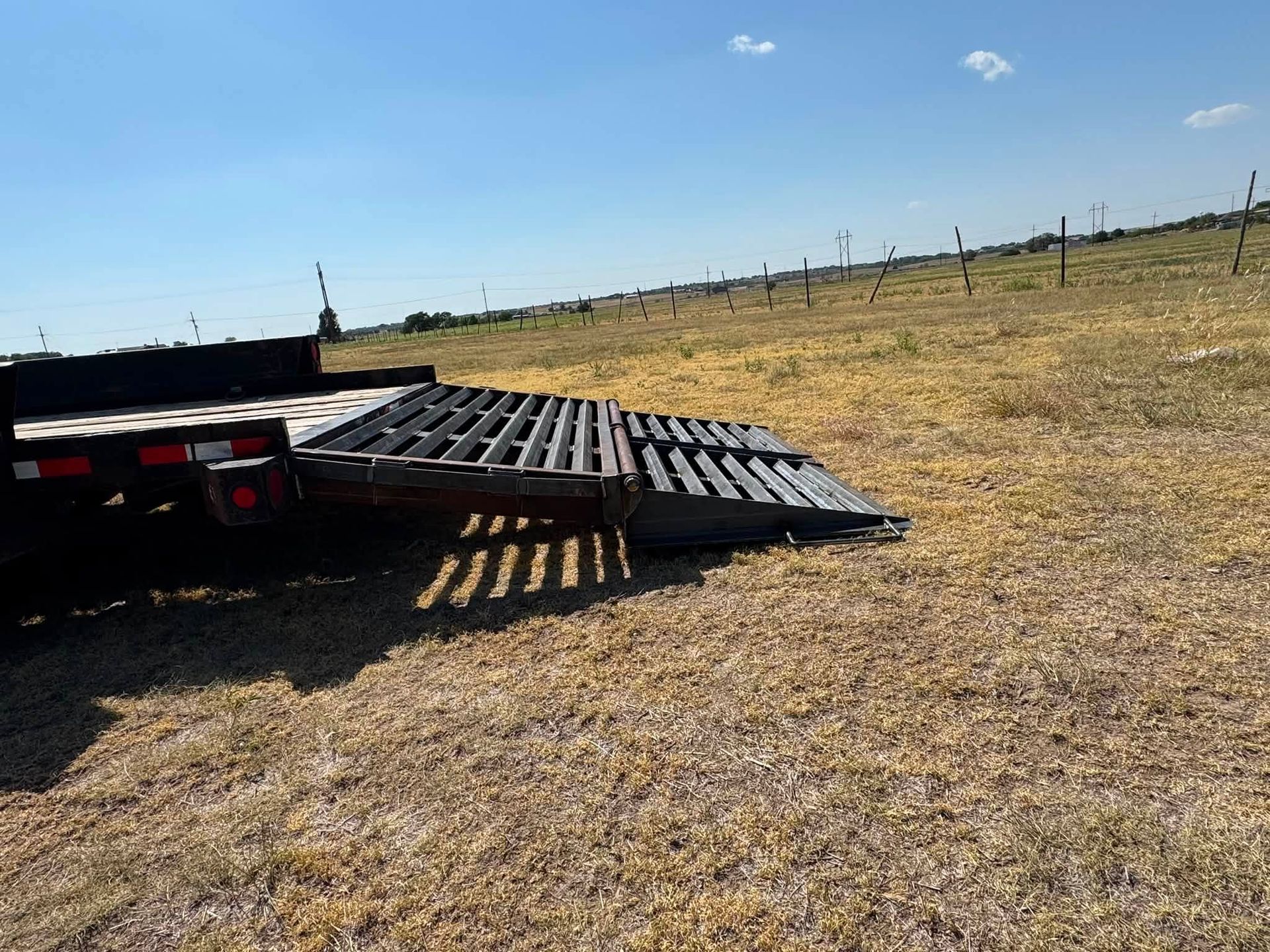 Flatbed trailer with a metal ramp in a dry grassy field under a clear blue sky