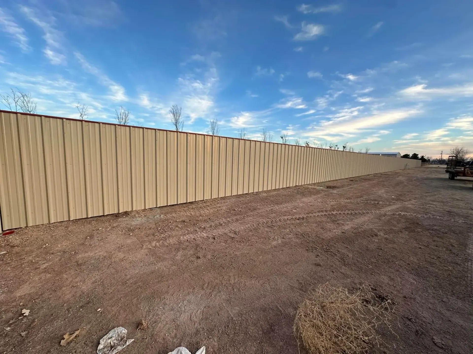 Empty dirt lot beside a tan wooden fence under a blue, cloudy sky