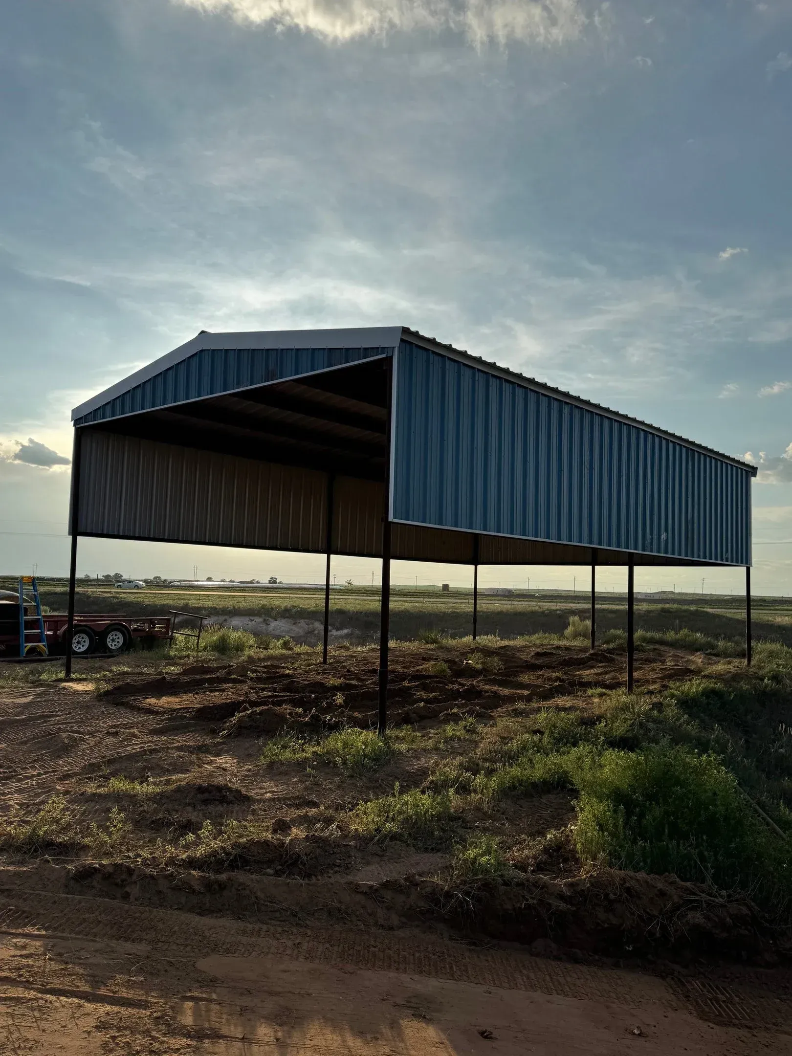 Blue corrugated metal pavilion on a dirt field at sunset, supported by thin posts.