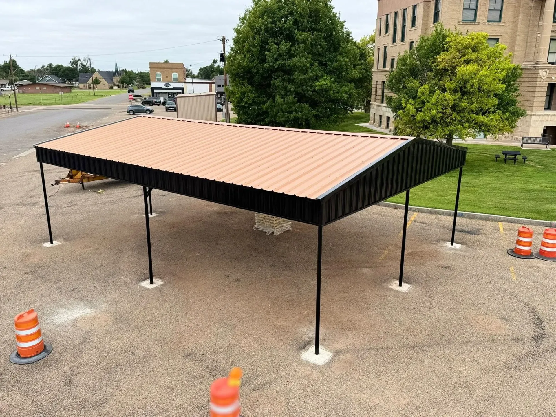 Temporary metal shelter with a slanted tan roof on a gravel lot near a building and road