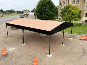 Temporary canopy tent with a tan roof and black frame set up on gravel beside a road and building.