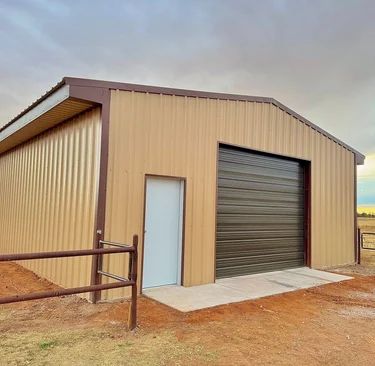 Tan metal storage shed with a large roll-up door and white side door on a dirt lot