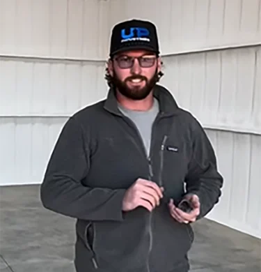 Man wearing a black cap and glasses stands indoors, holding a small device in a gray jacket.