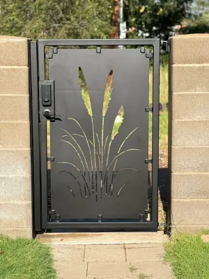 Black metal gate with a frosted panel and leaf design set in a stone wall, with a paved walkway in front.