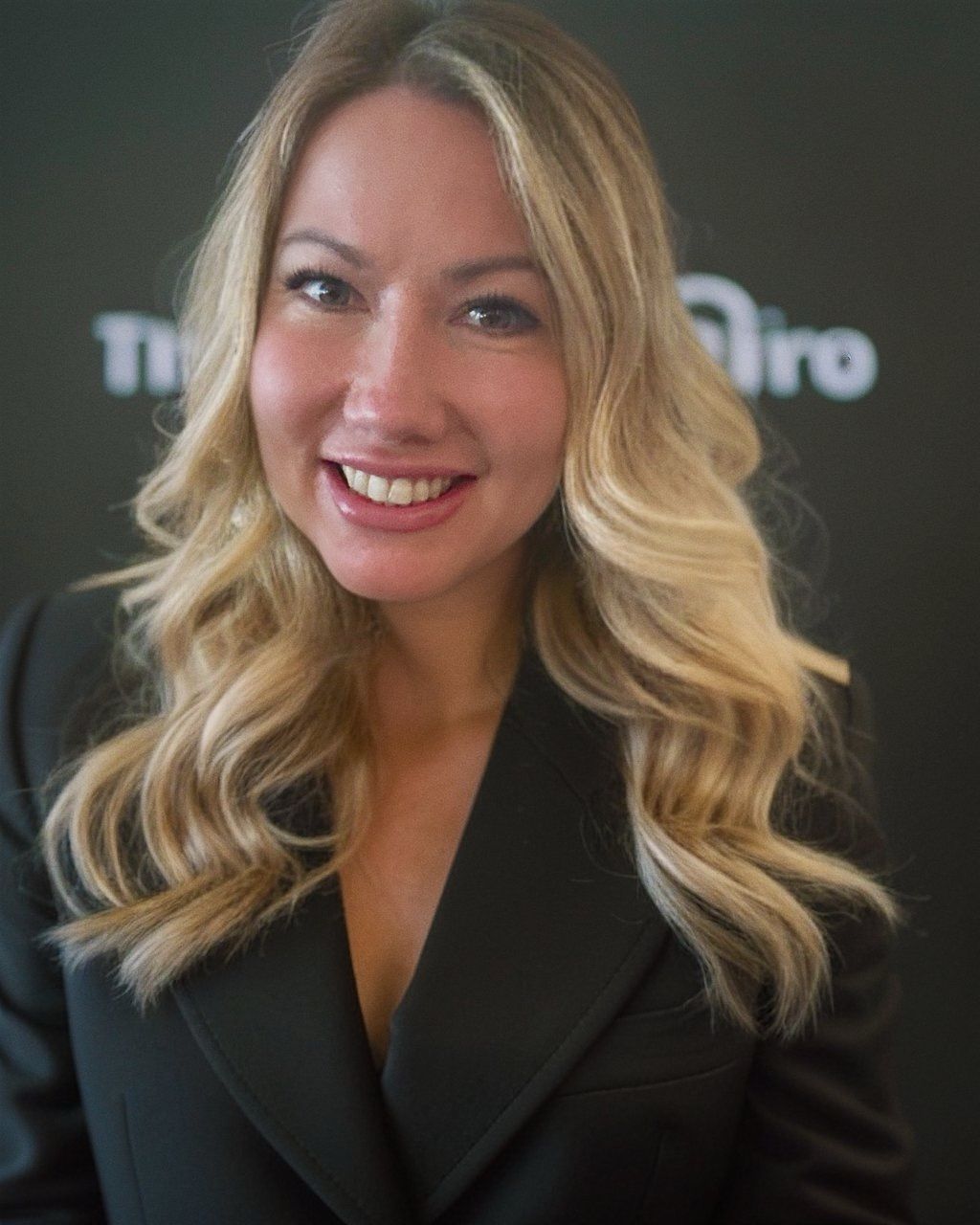 Smiling blonde woman in a black blazer against a dark backdrop