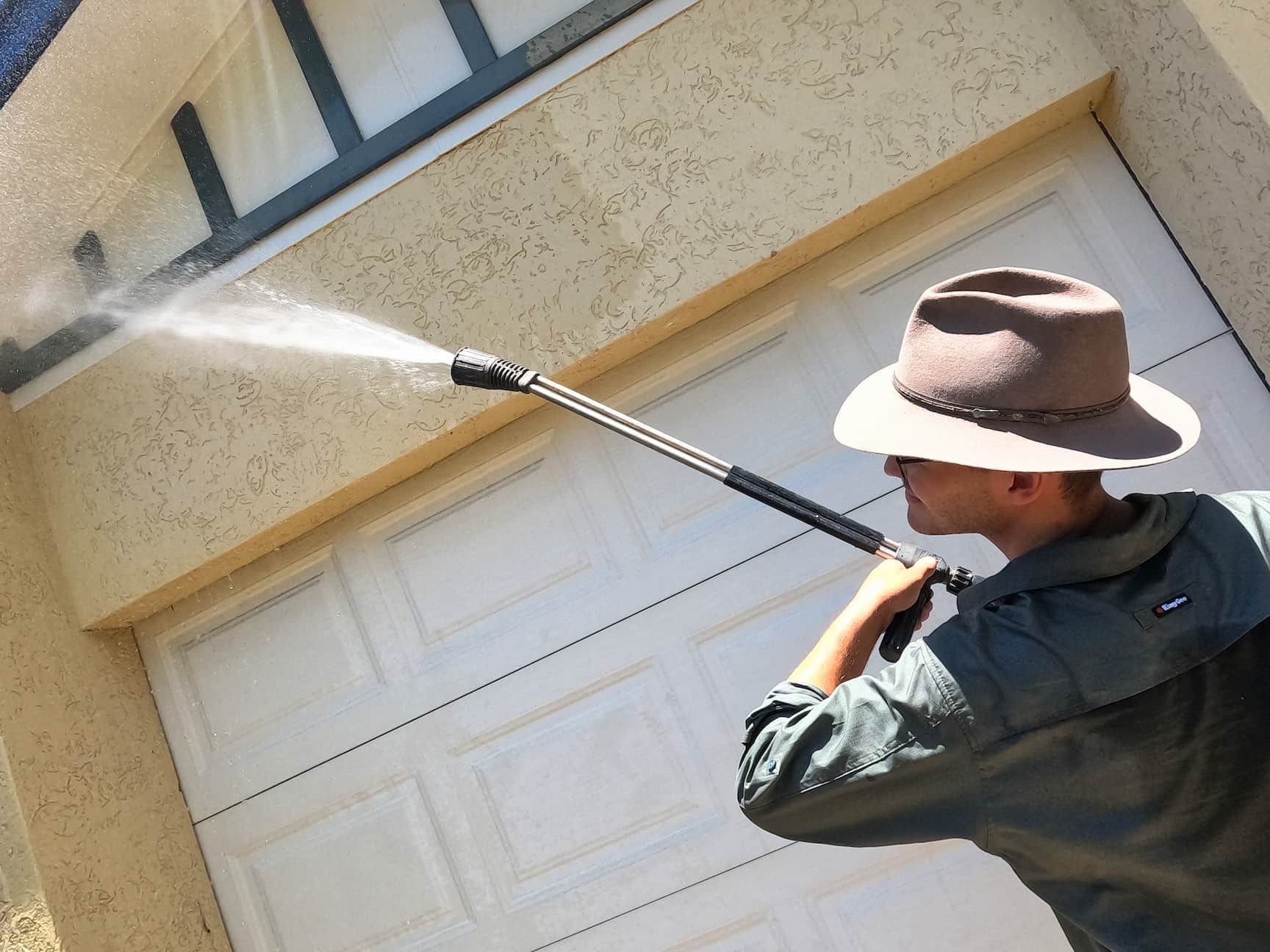 A Man in A Hat Using a High Pressure Washer to Clean a Garage Door — Cairns Bin Cleaning In Parramatta Park, QLD