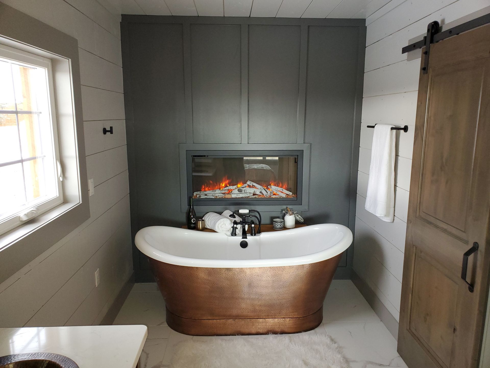 Modern bathroom with a copper soaking tub, fireplace, gray cabinets, and a wooden door