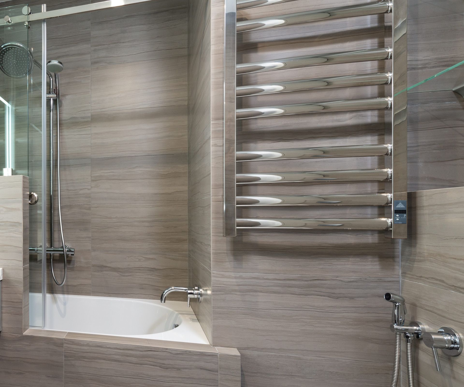 Modern bathroom with glass shower, bathtub, and chrome towel radiator on beige tile walls