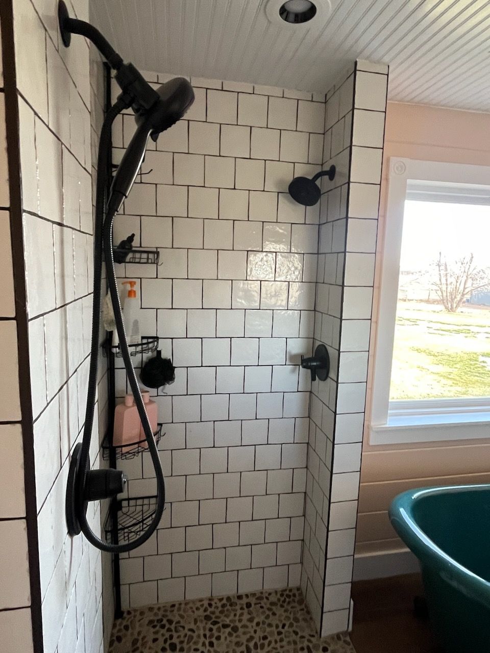 White-tiled bathroom shower with black fixtures and a window beside a bathtub.