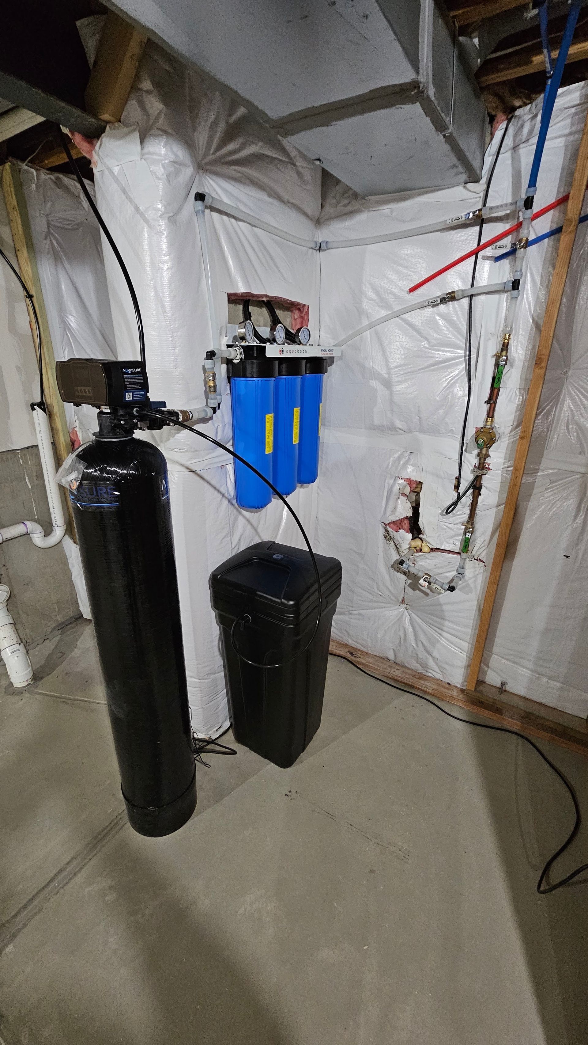 Water softener system in a basement, with black tanks, blue filters, and exposed ductwork and pipes.