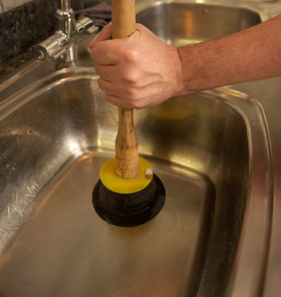 Hand gripping a wooden plunger over a kitchen sink drain with yellow and black rubber cup