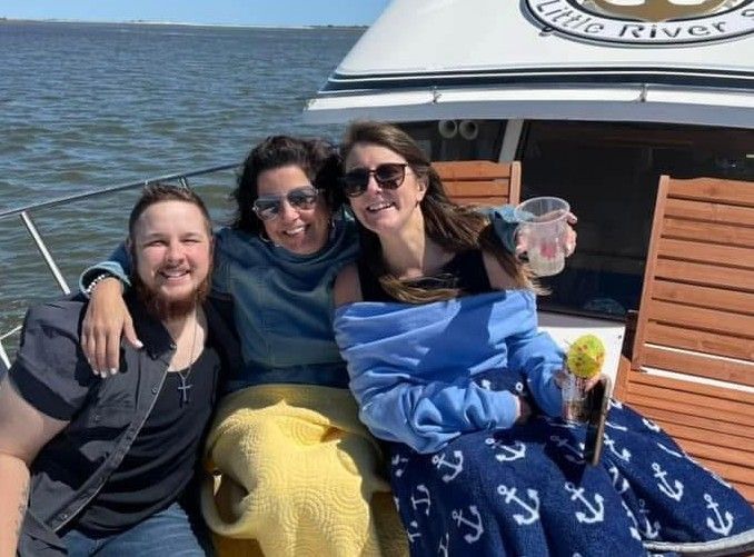 Three friends smiling on a boat, wrapped in blankets with drinks, on a sunny lake.