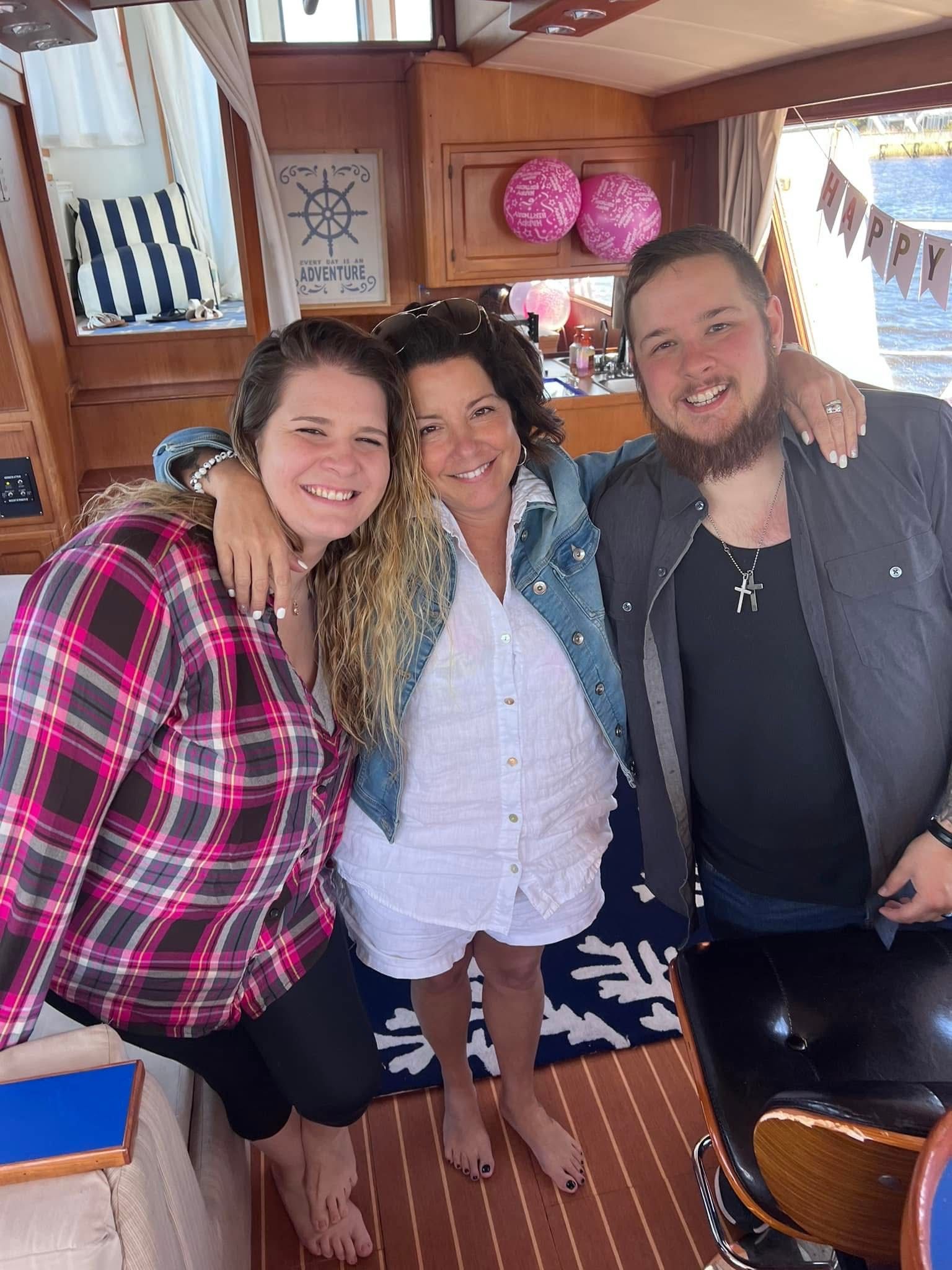 Three smiling people pose together in a wooden boat cabin, with striped curtains and pink decor.
