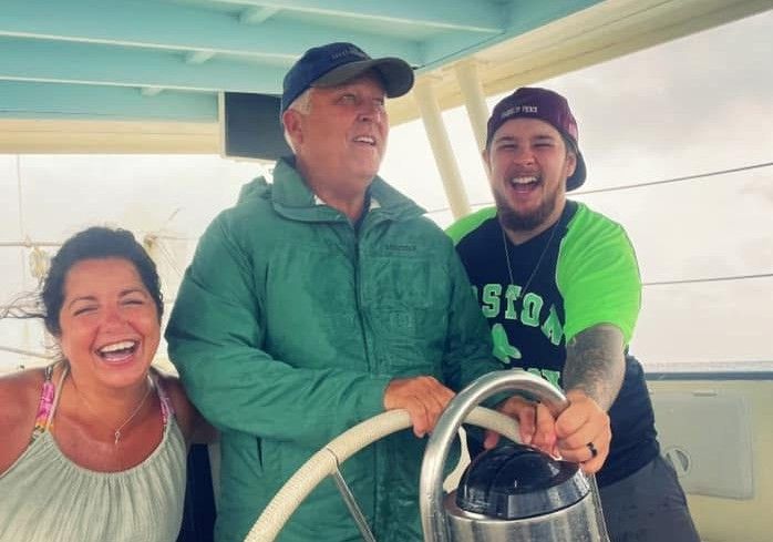Three people smiling on a boat, with one steering at the wheel under a blue canopy.