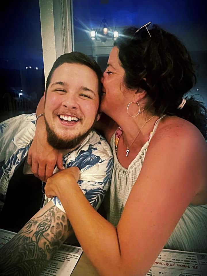 Two people smiling and embracing indoors, one kissing the other's cheek.