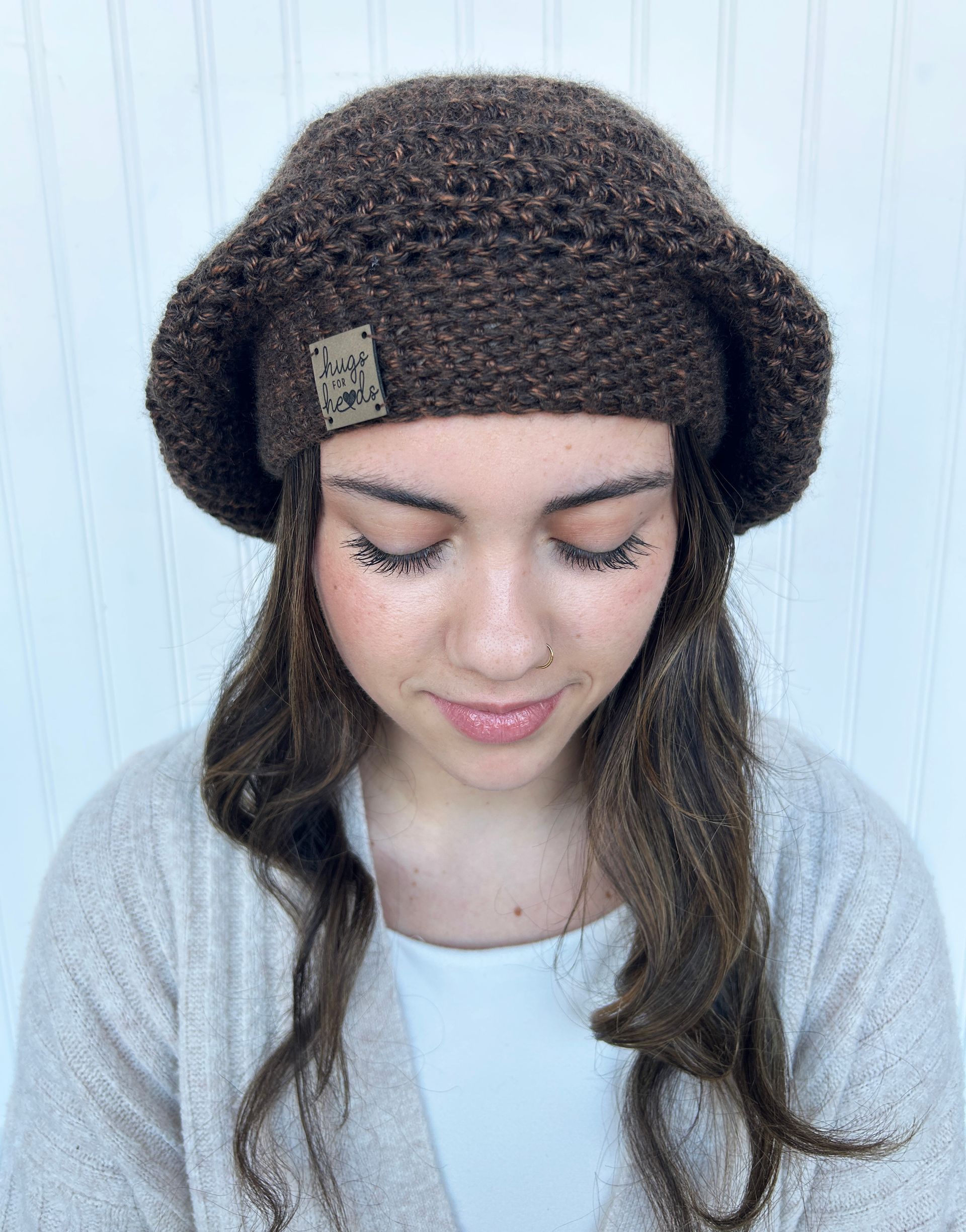Woman wearing a brown crocheted beret, looking down. Light-colored cardigan and white shirt. Against a white background.