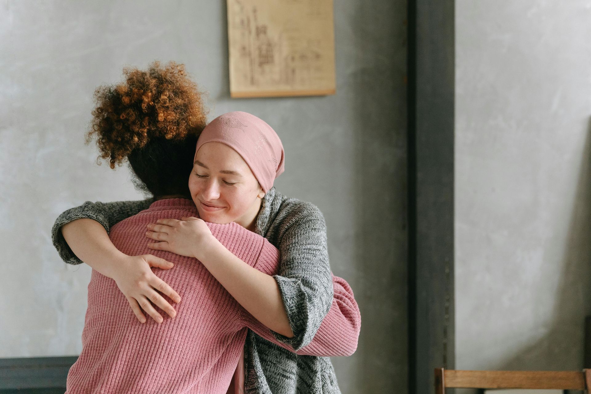 Two people embrace, one with a head covering, eyes closed, and smiling. Gray and pink clothing. Indoors.