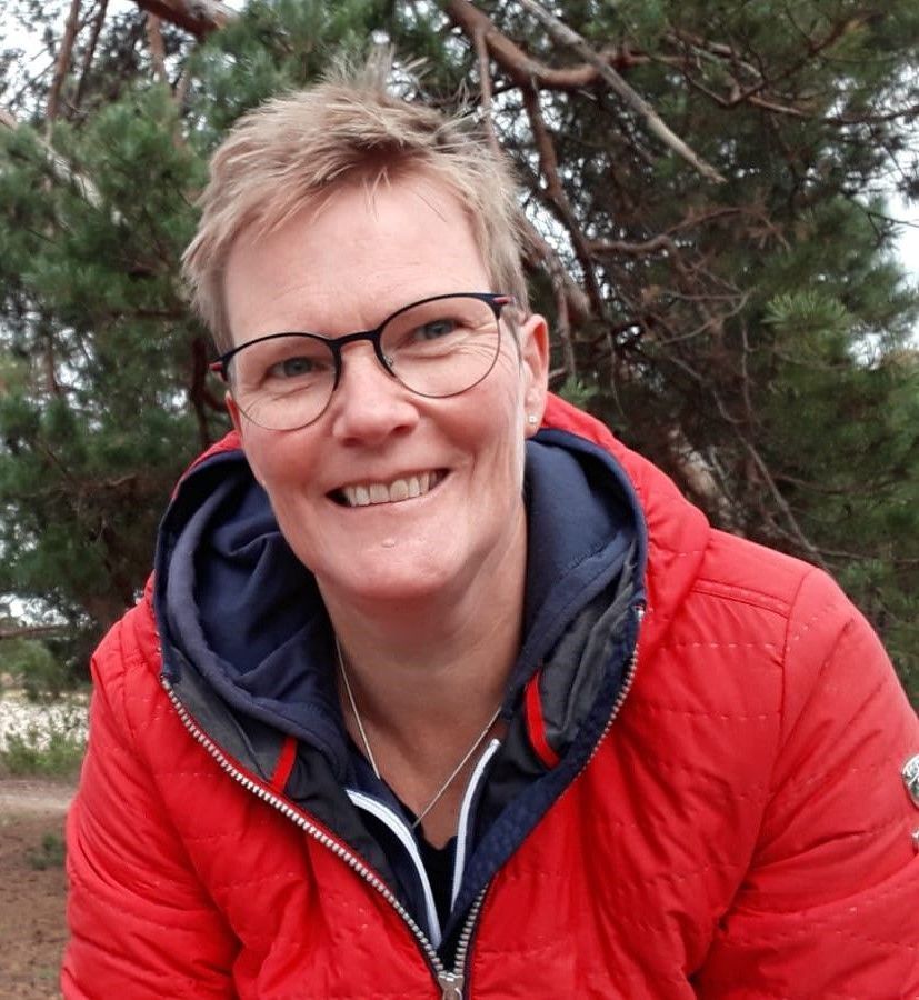 Een vrouw met een bril en een rood jasje lacht naar de camera.