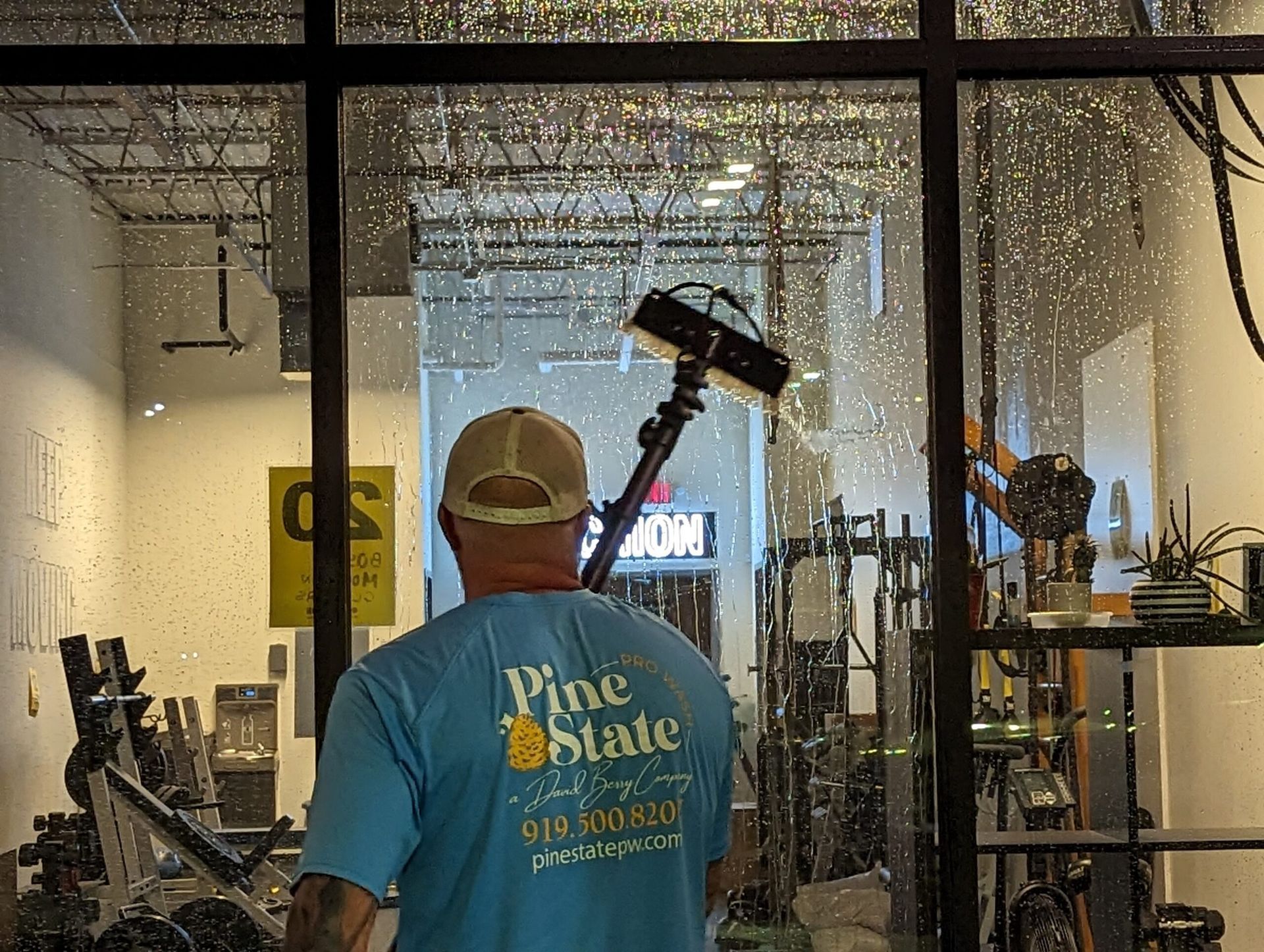 A worker wearing a blue Pine State t-shirt uses a pole to clean a large glass window of a gym from the outside.