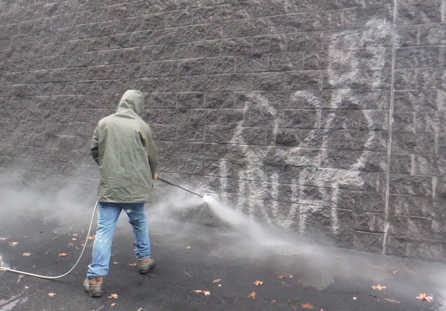 A person in a hooded jacket uses a power washer to remove graffiti from a stone wall.