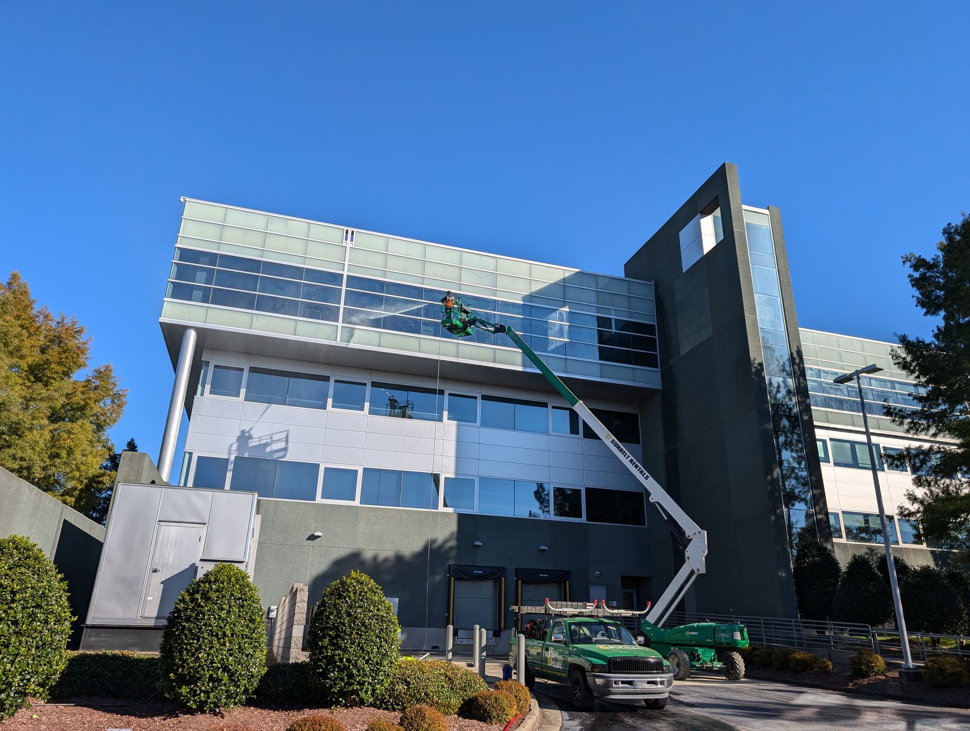 A green aerial lift reaches toward the exterior windows of a grey office building on a clear, sunny day.