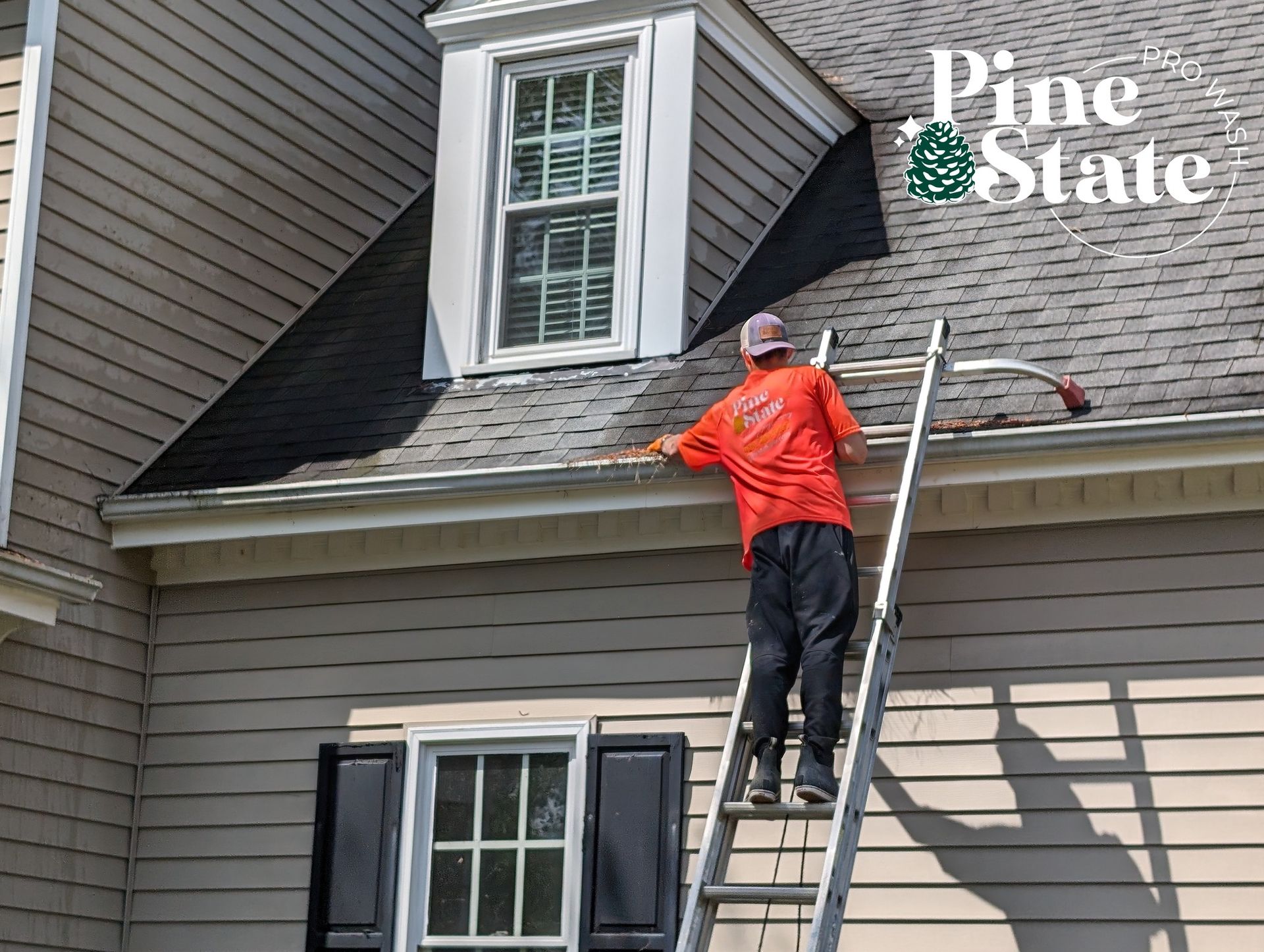 A worker in an orange shirt on a ladder cleaning a roof gutter on the side of a tan house.