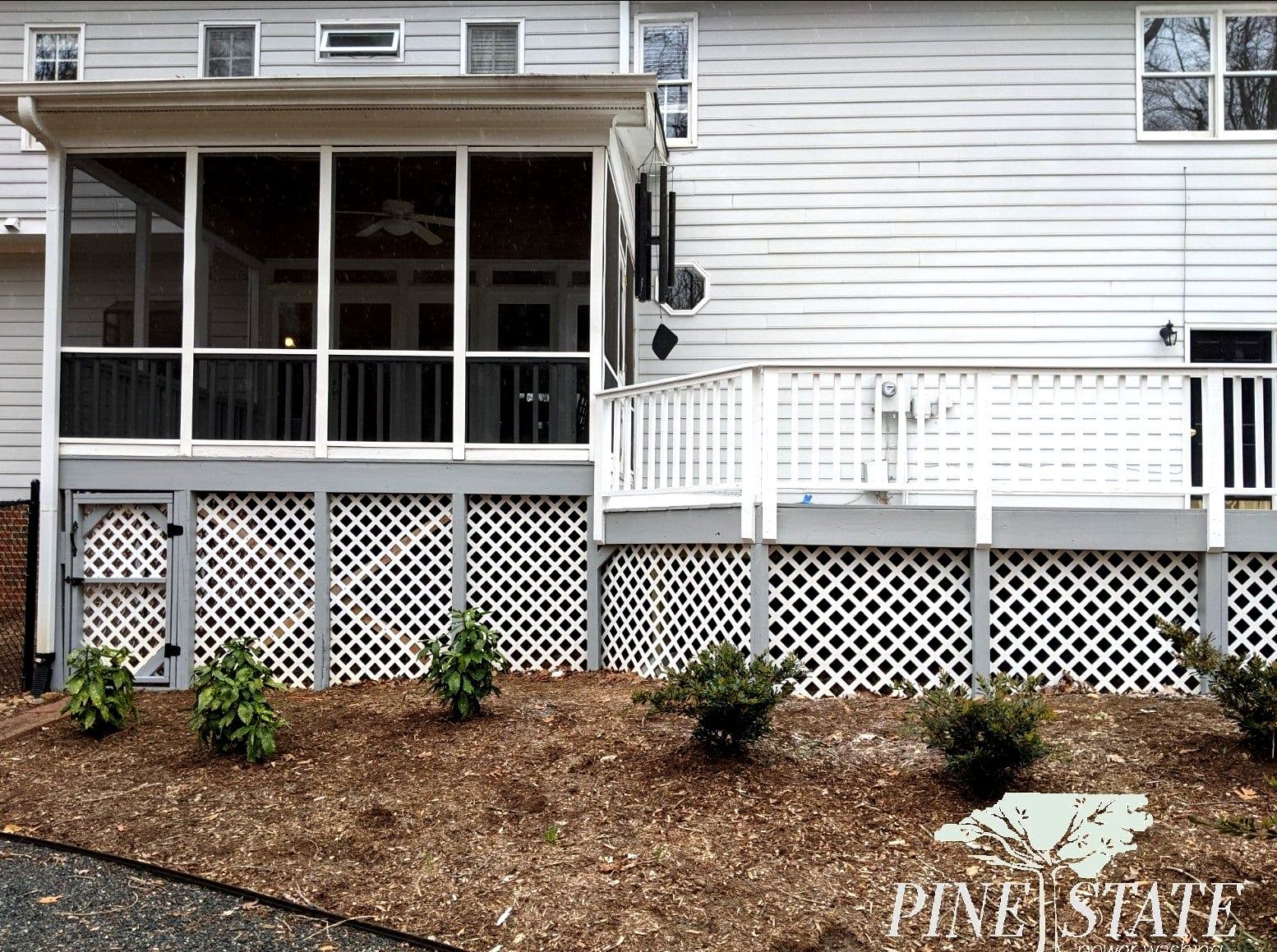 A gray house exterior featuring a screened-in porch, a wooden deck, and white lattice skirting over a mulched landscape.