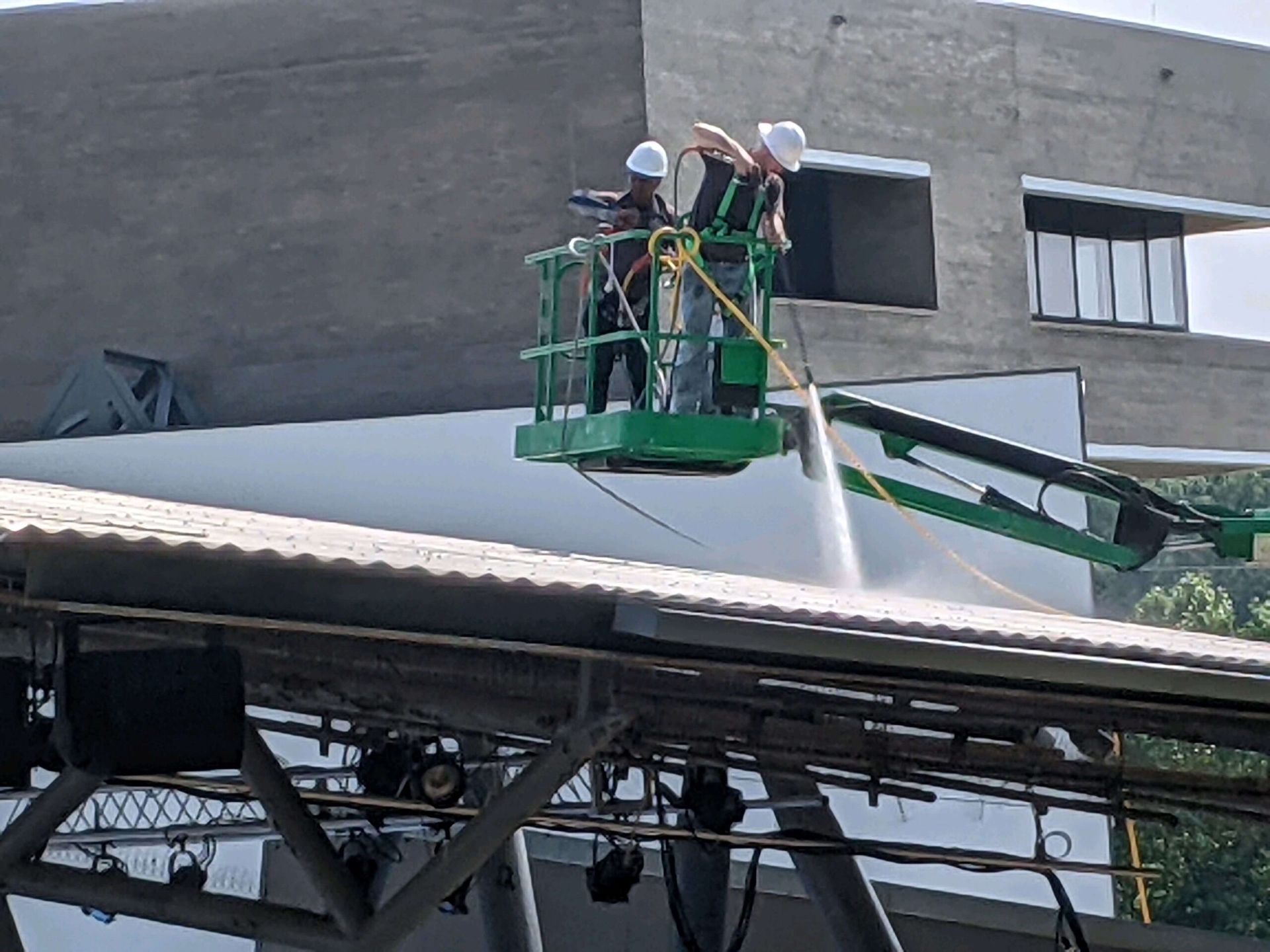 Two workers in hard hats use a pressure washer to clean a white roof surface from an aerial lift.