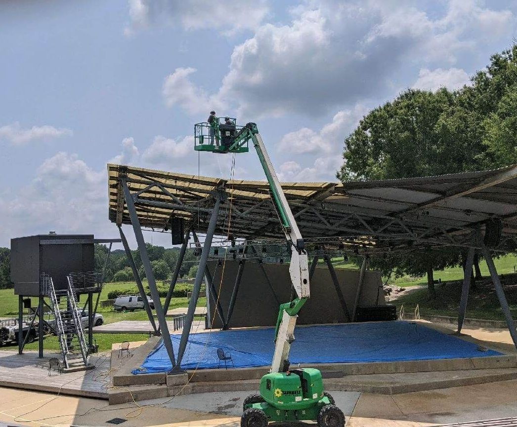 A worker in a boom lift reaches the roof of an outdoor stage structure under a blue sky.
