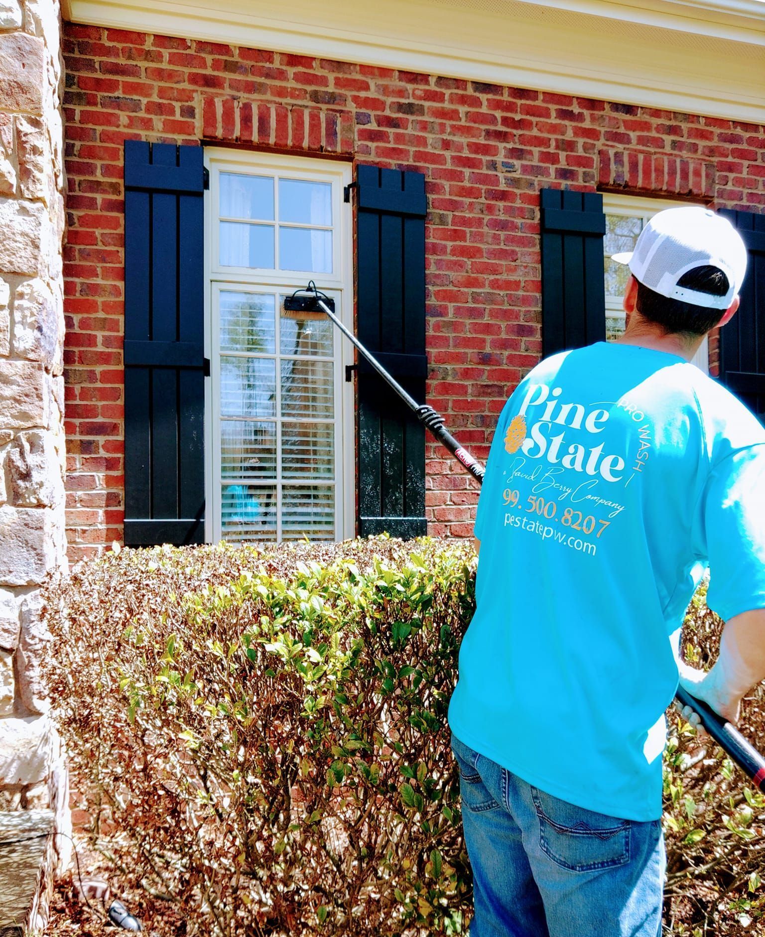 A person in a blue Pine State t-shirt and white cap uses an extension pole to clean a window on a brick house.