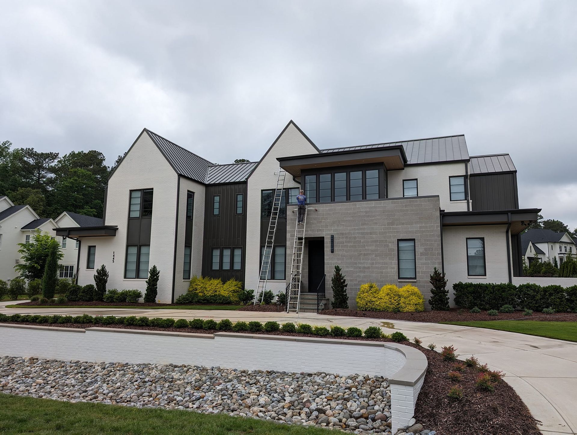 A modern two-story home with white and stone siding, dark accents, and a metal roof under a cloudy sky.