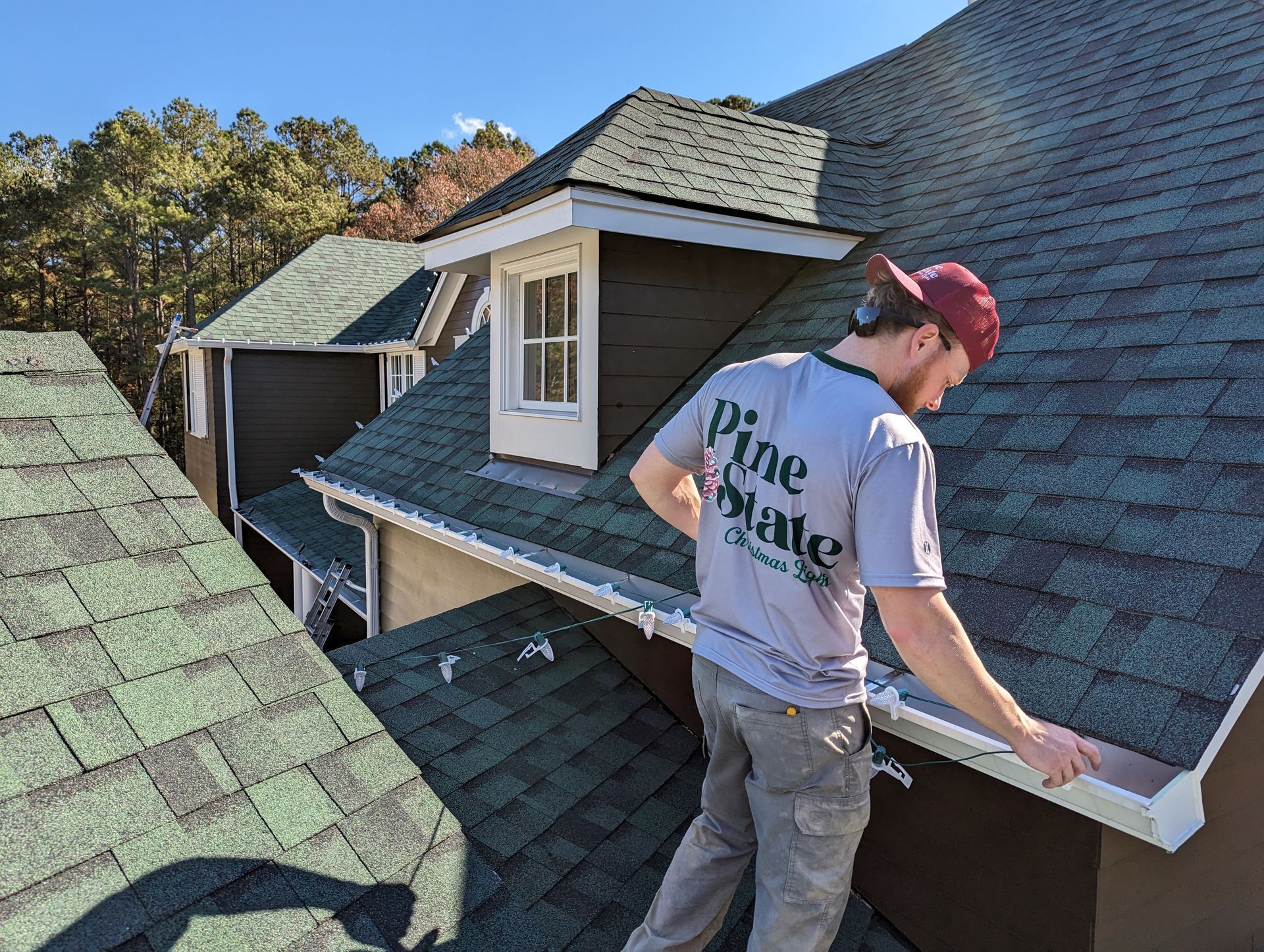 A worker wearing a Pine State shirt installs white gutter trim on a shingled roof next to a dormer window.