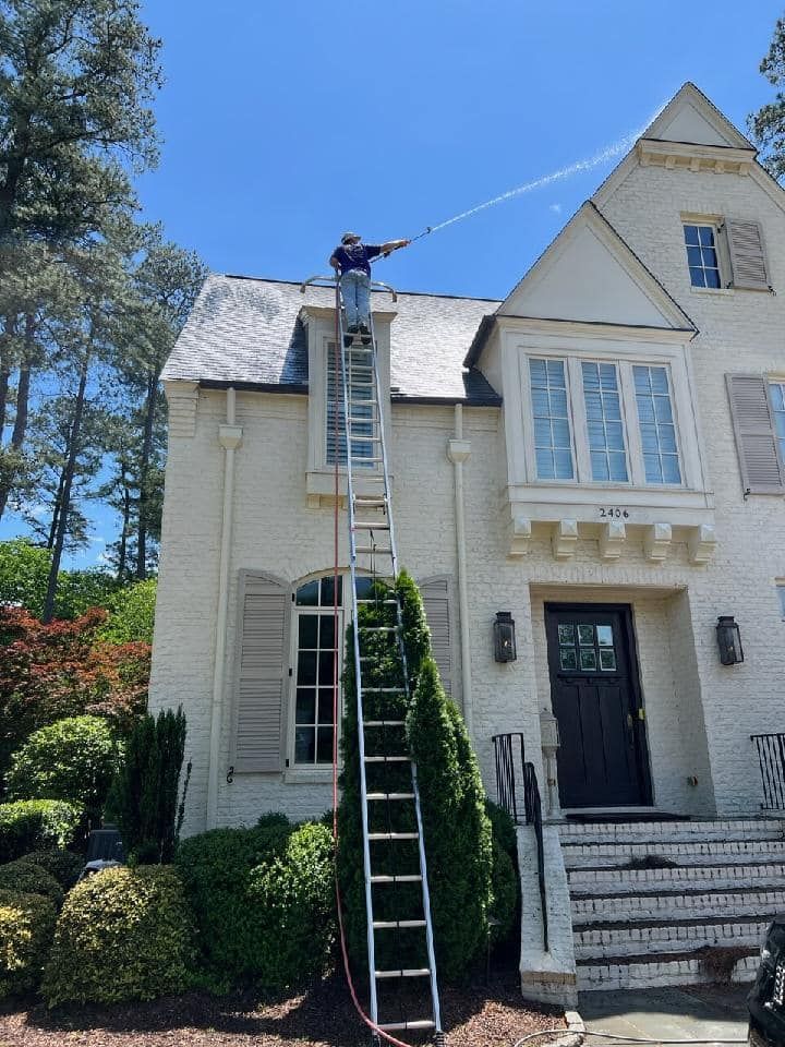 A man on a ladder is spraying water on the roof of a white house.