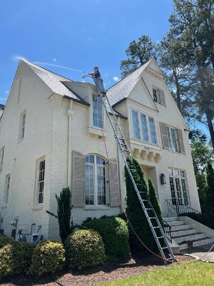 A man on a ladder is cleaning the roof of a white house.