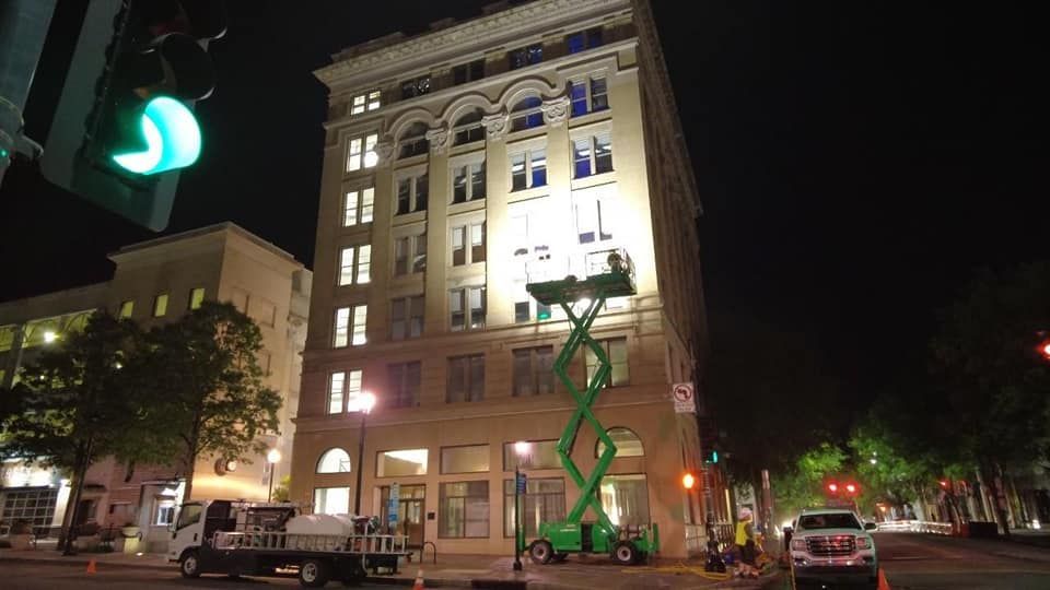 Nighttime view of a lit-up historic building with a green scissor lift parked in front, with vehicles and trees nearby.