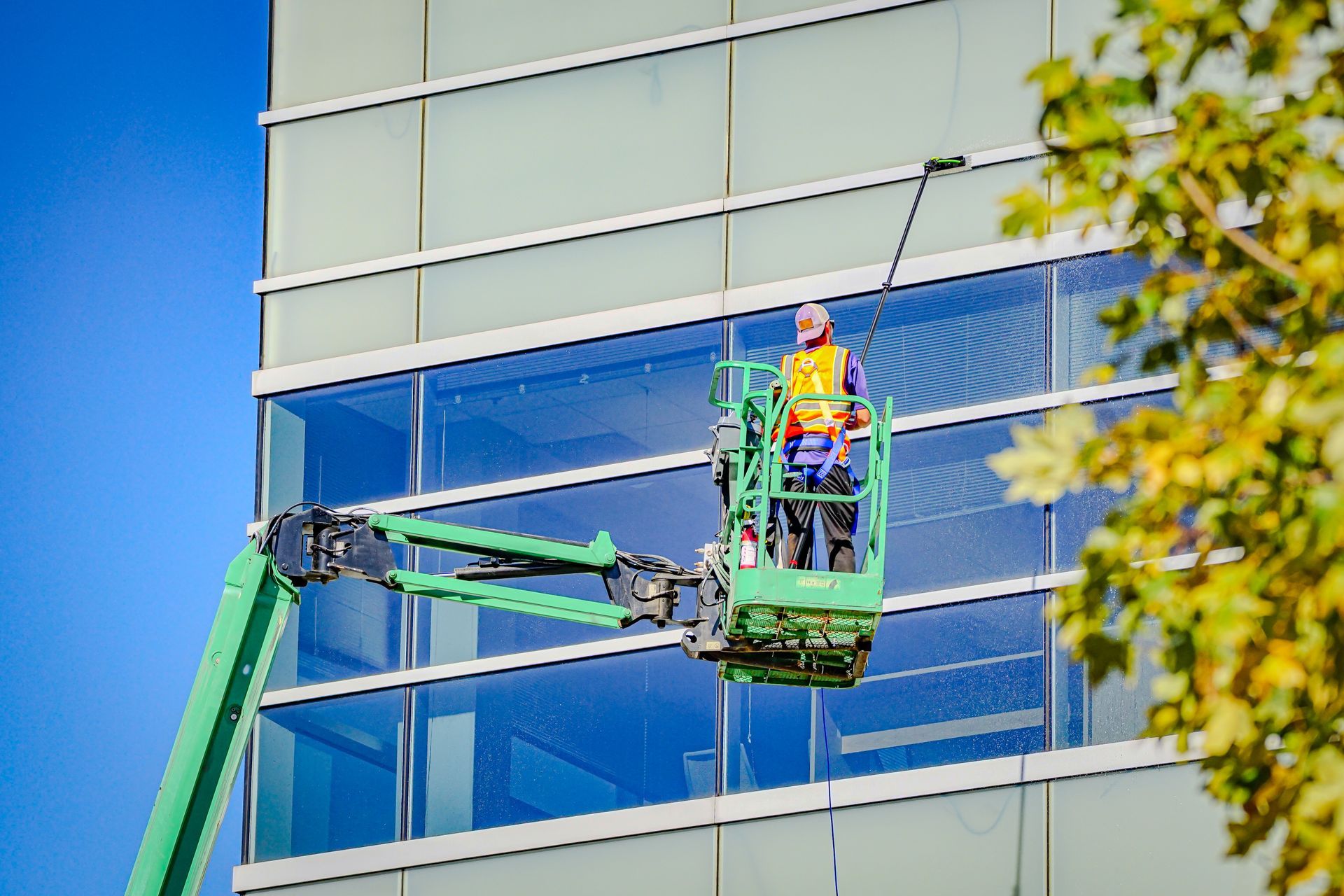 A worker in a bright yellow safety vest cleans the glass facade of a modern building from a green aerial lift platform.
