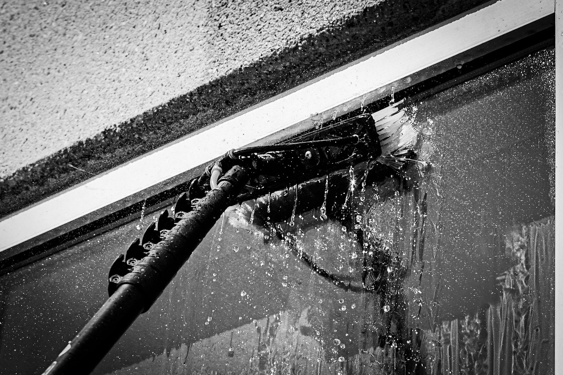 A close-up, black-and-white view of a brush on a long pole spraying water to clean a glass surface.