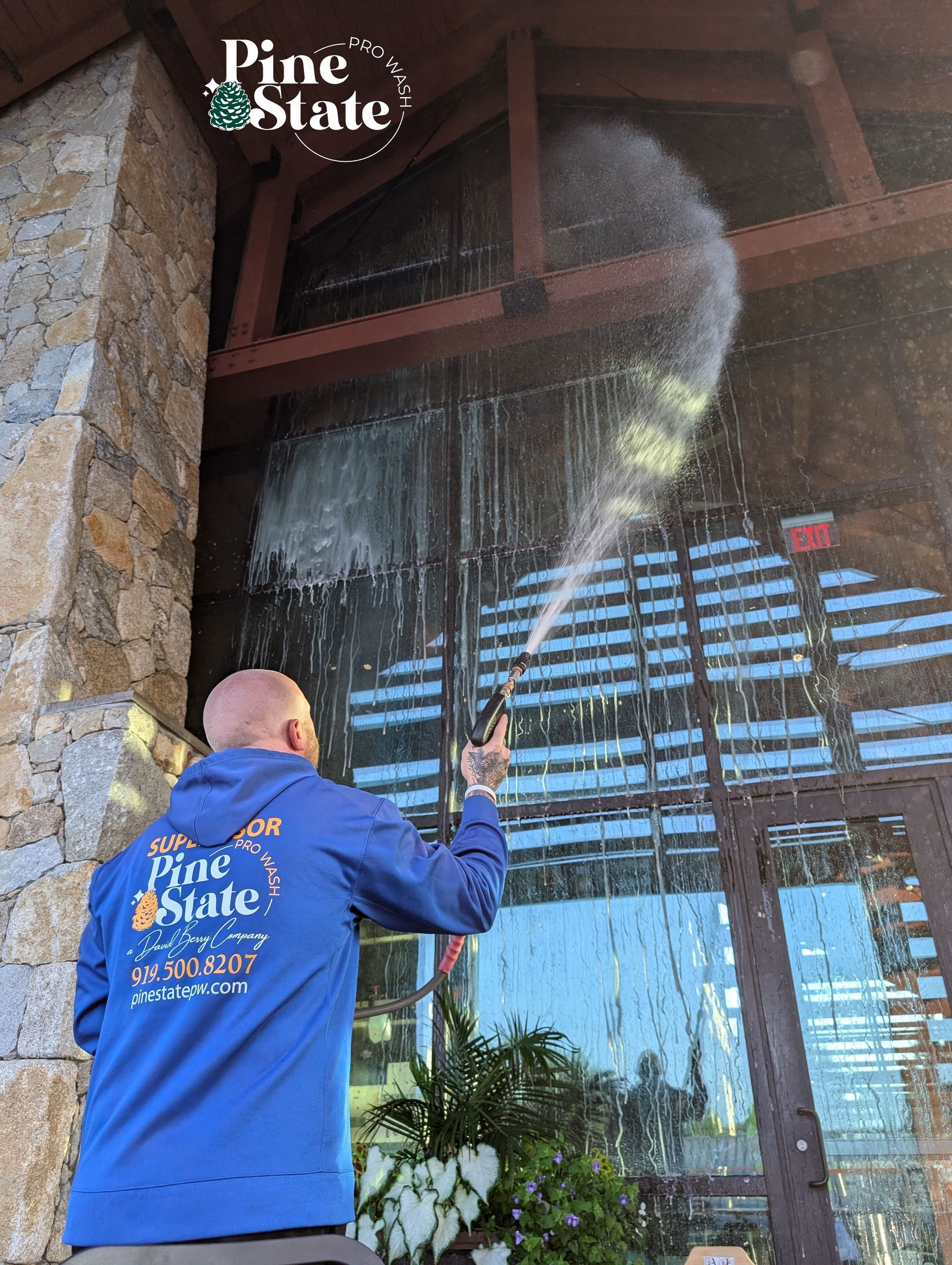 A person wearing a blue Pine State hoodie pressure washes the glass facade of a stone building.