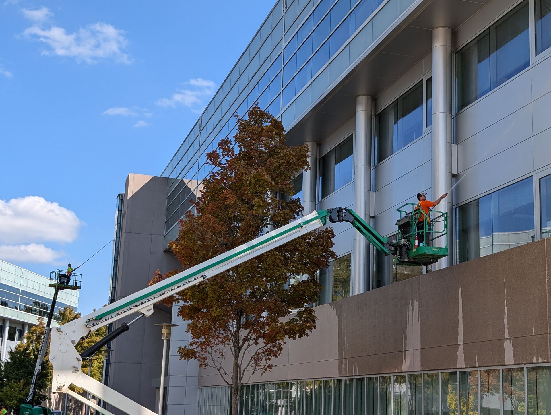 A worker in a bucket lift cleans the exterior windows of a modern office building on a sunny day.