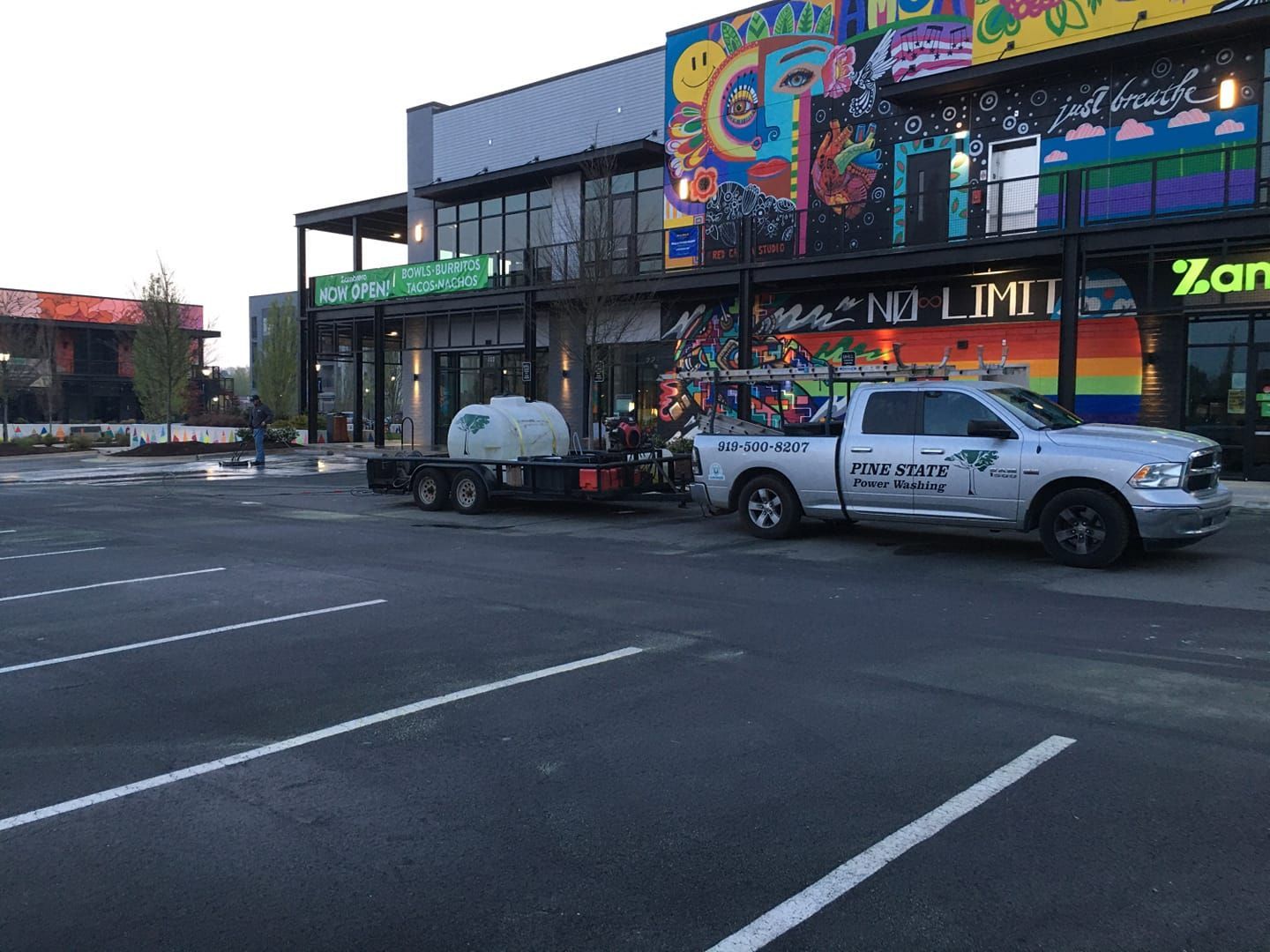 A silver truck towing a water tank trailer is parked in an asphalt parking lot in front of a modern building with murals.