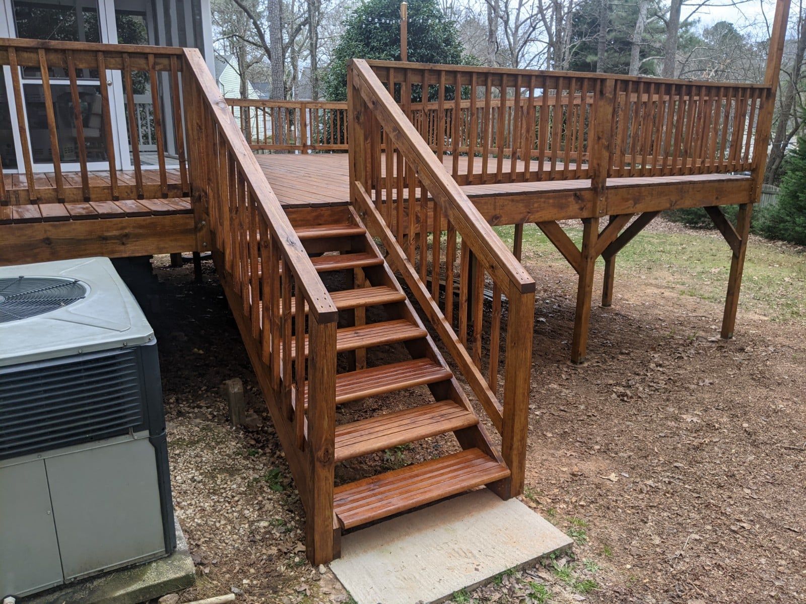 A set of wooden stairs with railings leading up to a raised outdoor deck next to an HVAC unit in a wooded yard.
