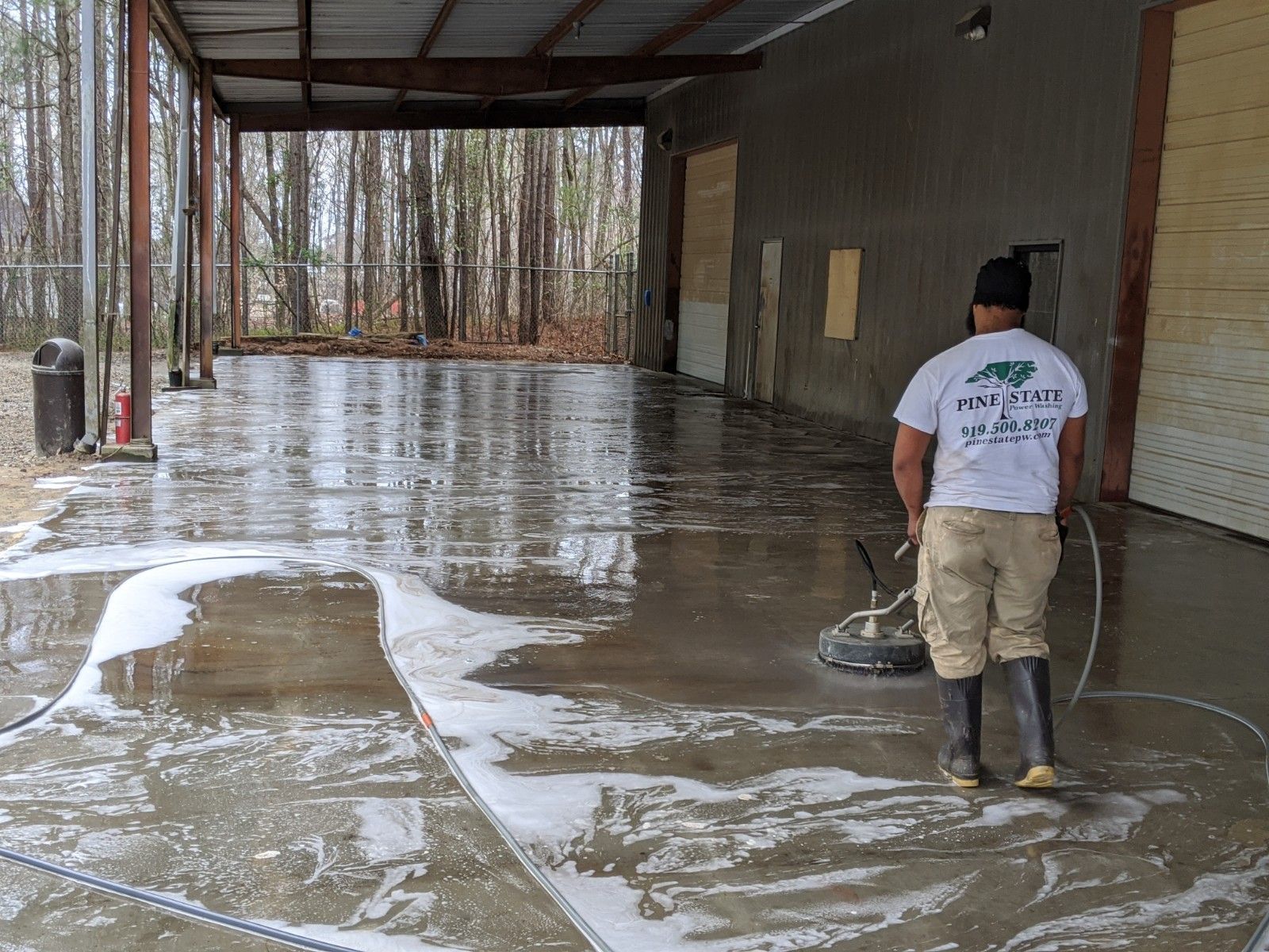 A person uses a circular power washer to clean a wet concrete floor under a covered outdoor structure.
