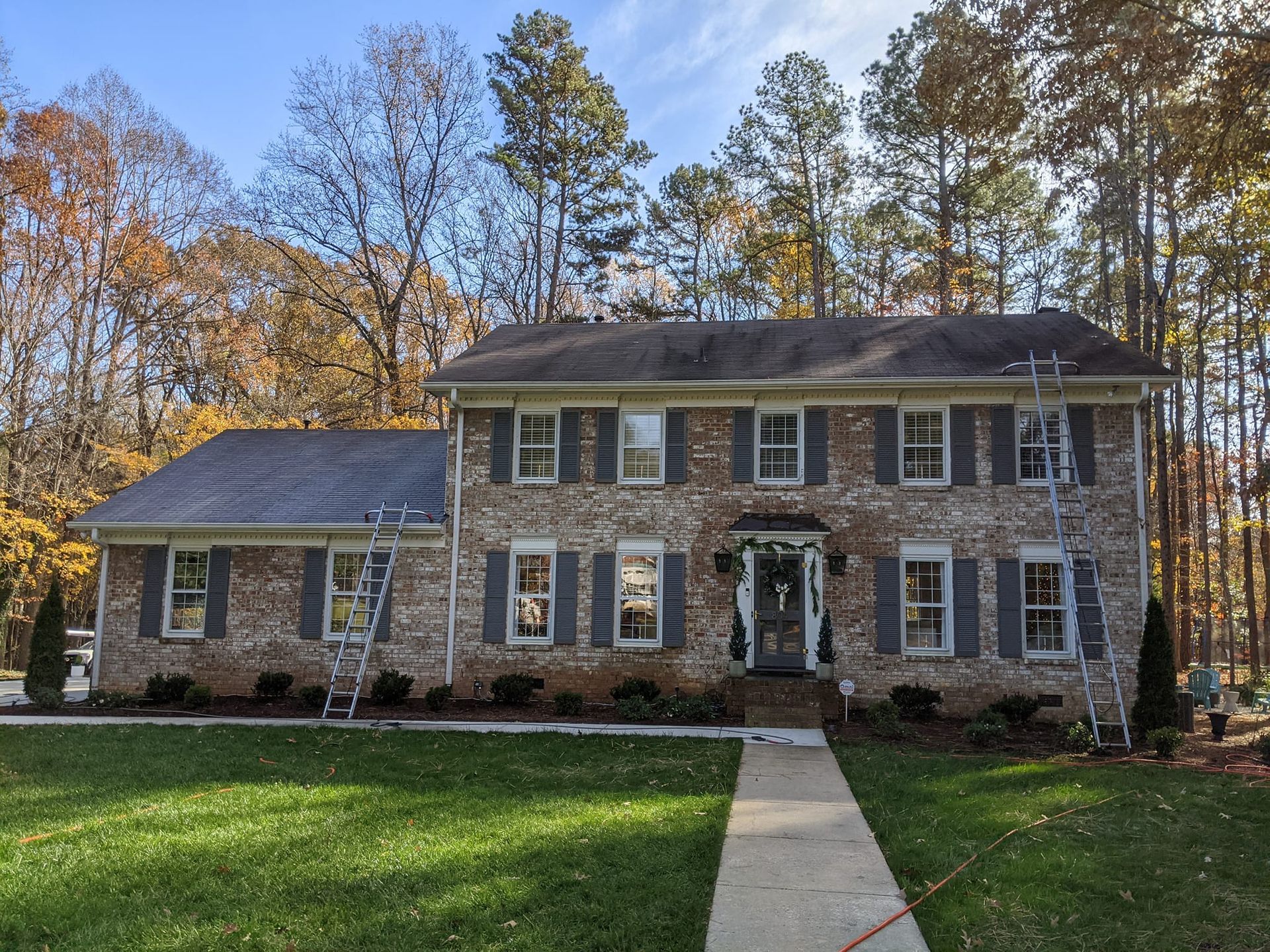 Two-story brick house with a slate roof, gray shutters, and a front walkway, set against a backdrop of autumn trees.